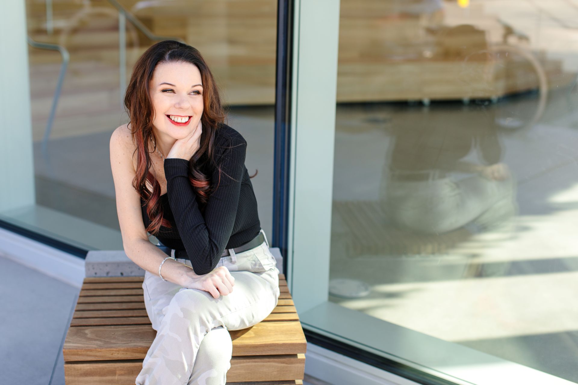 A woman with long brown hair in a black top and light pants sits on a wooden bench outside, smiling with her hand resting on her chin.